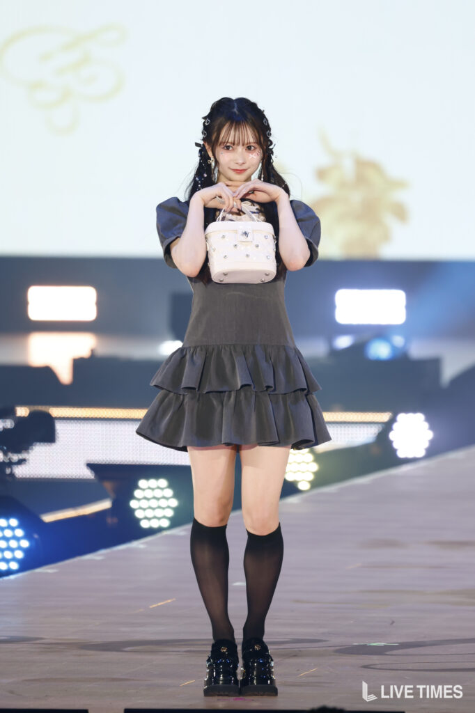 Young woman on stage posing with a small studded white handbag, wearing a dark ruffled dress and knee-high socks.
