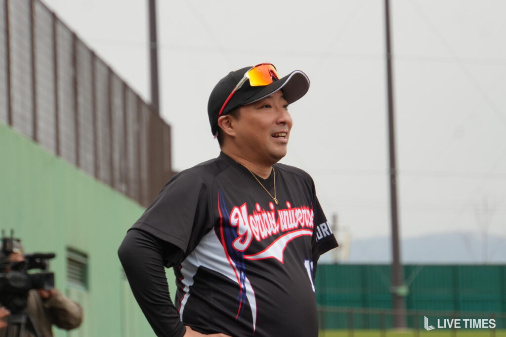 Man in a black athletic jersey standing on a baseball field, smiling, wearing a cap with sunglasses on top.