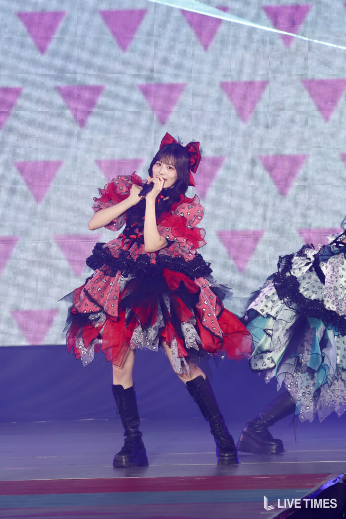 Singer performing on stage wearing a red and black layered dress with a large bow, holding a microphone.