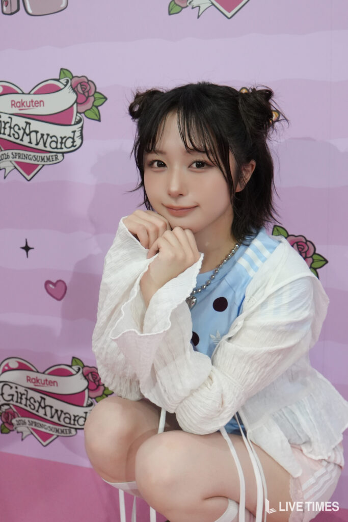 Young woman with dark hair styled in two buns, wearing a light blue polka-dot dress and white sheer cardigan, posing with hands near her chin in front of a pink backdrop with event graphics.