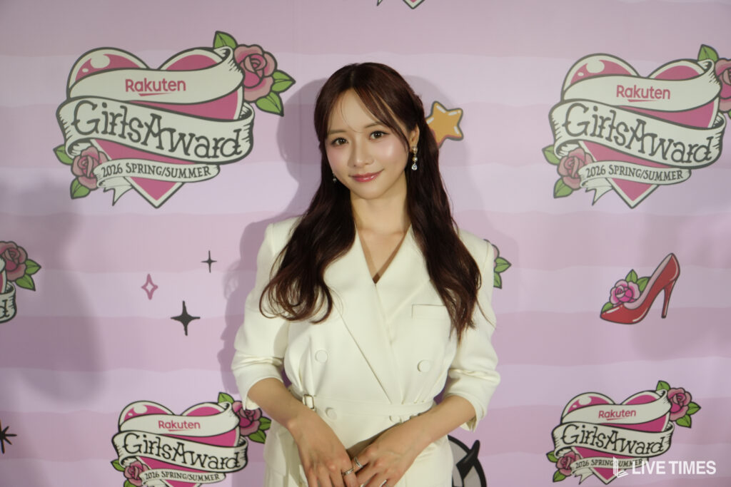 Woman in a white blazer posing in front of a pink Rakuten GirlsAward backdrop with logos and roses.