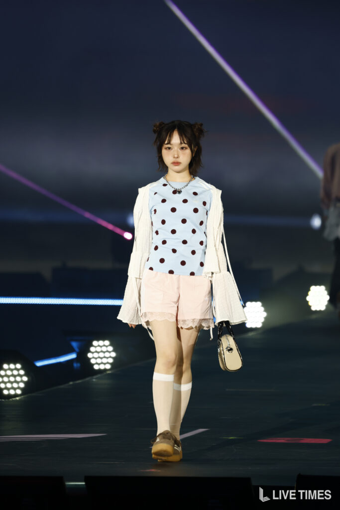 Model walking a runway at night in a light blue polka-dot top, pale pink shorts, white cardigan, brown shoes, and a beige handbag.