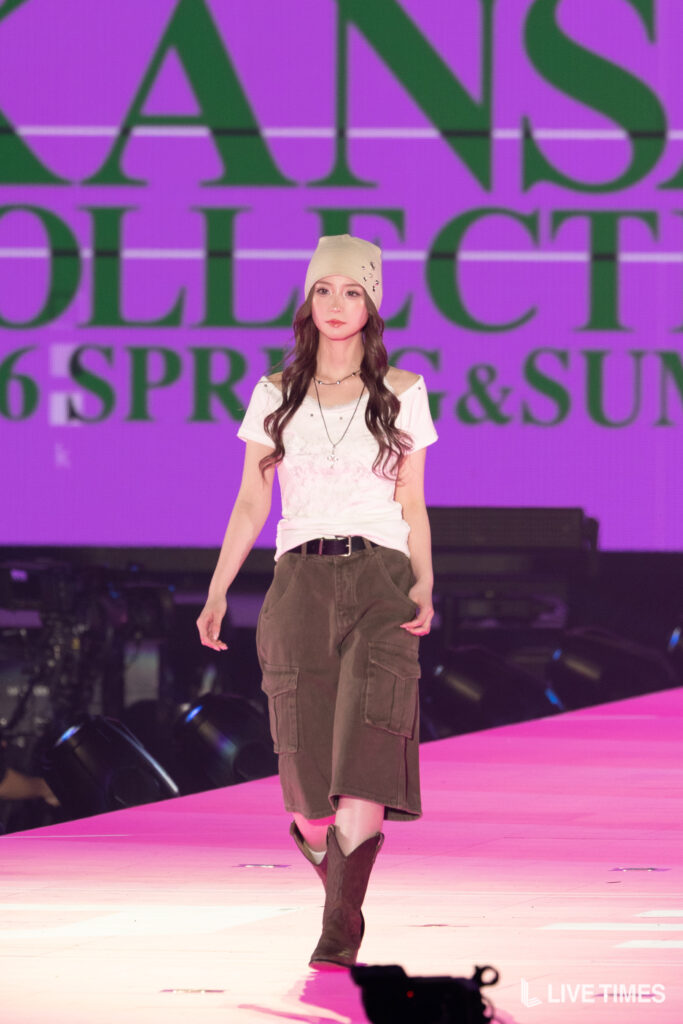 Female model walking on a pink runway wearing a beige beanie and olive cargo skirt, with a purple backdrop behind her.