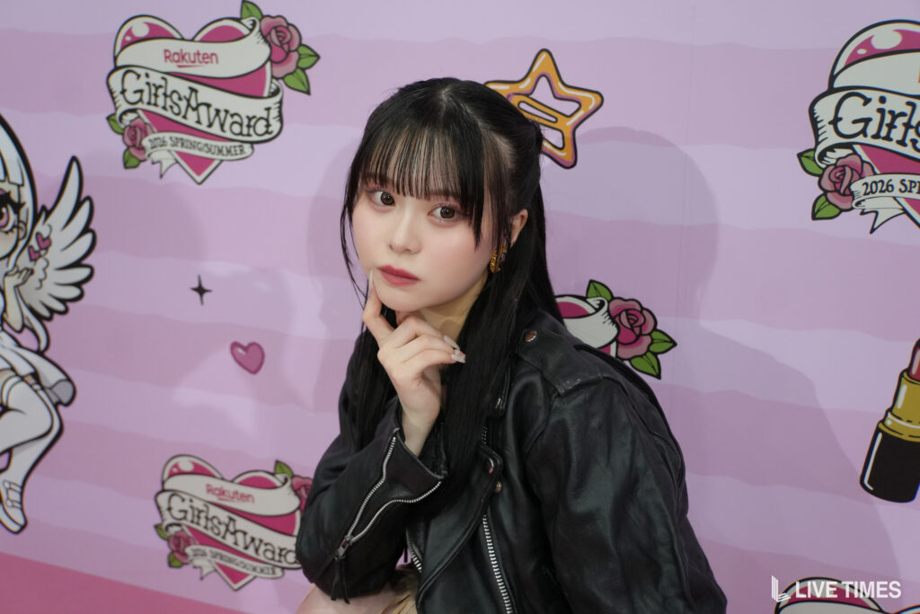 Portrait of a young woman with long black hair and bangs, wearing a black leather jacket, posing with her hand on her chin against a pink illustrated backdrop with 'GirlsAward' branding.