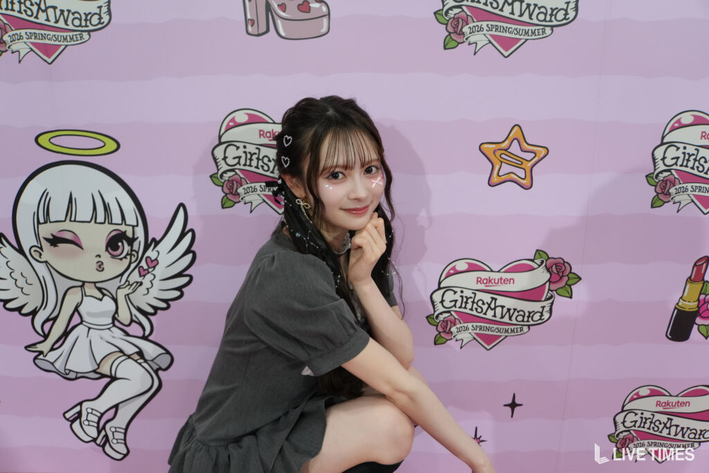 Young woman posing with hands under chin in front of a pink GirlsAward backdrop, wearing a gray dress and heart hair clips.