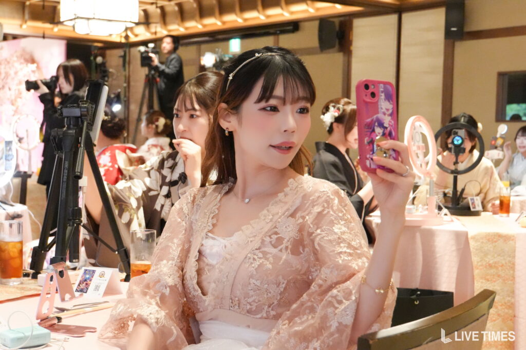 Woman in a pink lace dress takes a selfie with a pink phone at a makeup booth, with photographers and beauty products in the background.