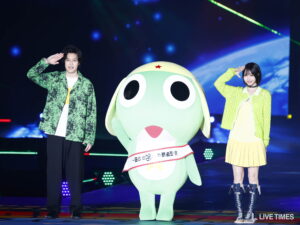 Mint-green mascot with a red star on its head and a sash, flanked by two performers saluting on a brightly lit stage.