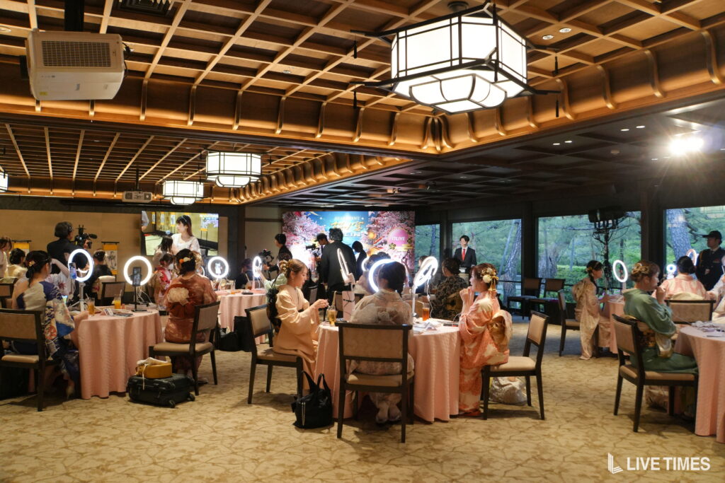 Banquet hall filled with guests in traditional kimonos seated at round pink-clothed tables under warm wooden ceiling lights, with a stage and large banners in the background.