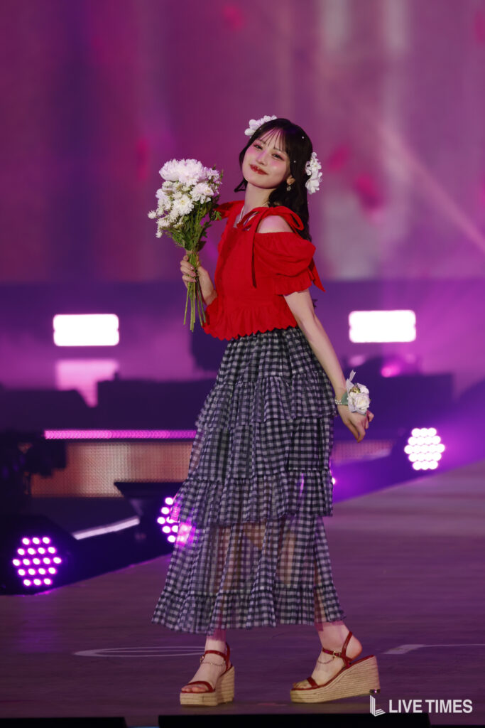 A young woman in a red off-shoulder top and tiered plaid skirt on stage, holding a bouquet of white flowers.
