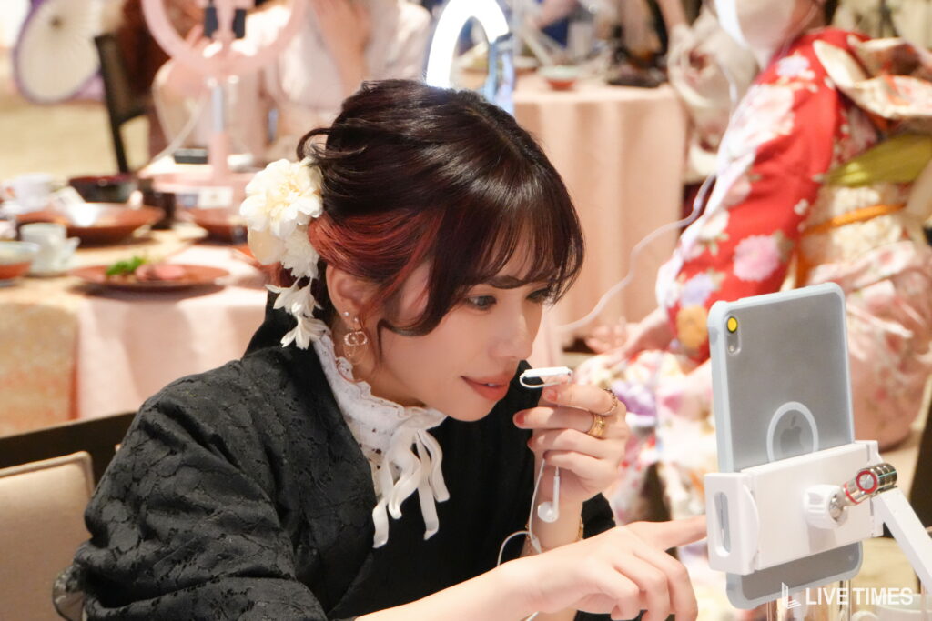 Woman with a white flower hair accessory looks at a smartphone mounted on a stand, likely livestreaming or video chatting, at a social event.