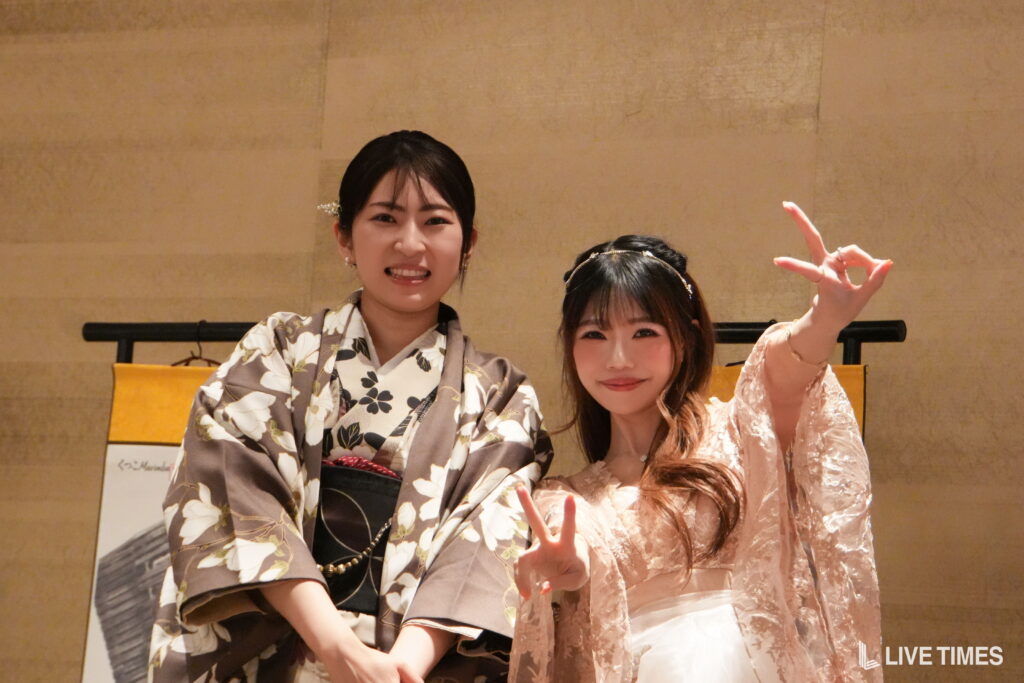 Two women in traditional kimonos posing and smiling at the camera in a beige hallway; one shows a peace sign with her hand.