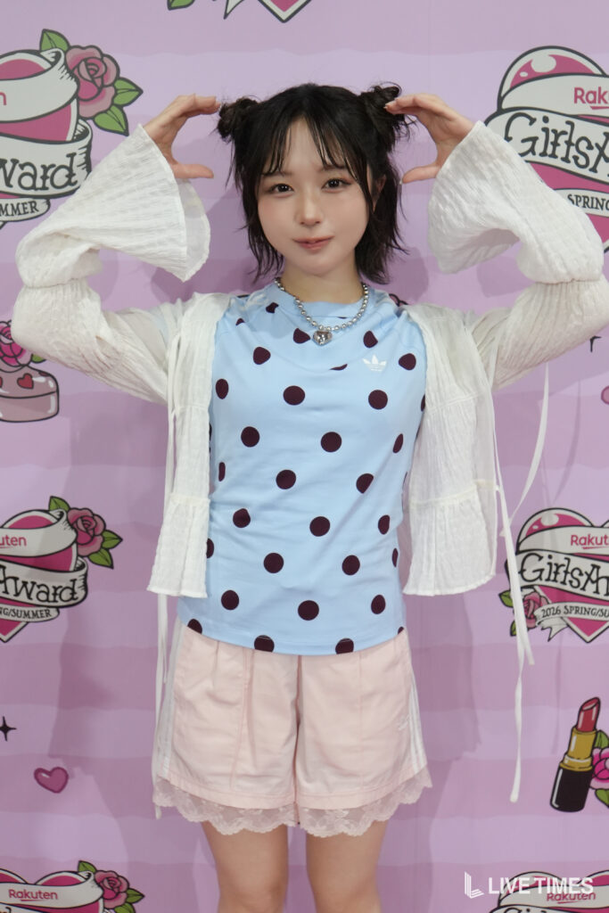 Woman posing with her hands shaped like bunny ears above her head, wearing a light blue polka-dot top and white cardigan against a pink backdrop.