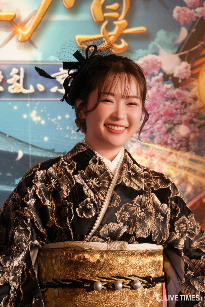 Smiling woman in a black and gold brocade kimono with an ornate hairpiece and pearl necklace, standing before a colorful backdrop.