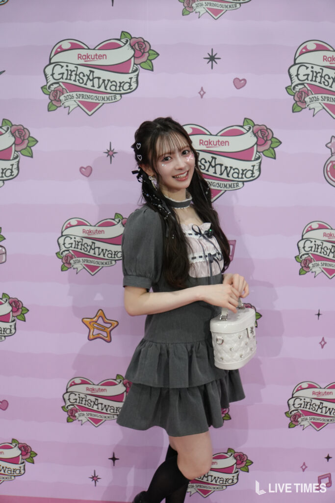 Young woman posing in front of a pink GirlsAward backdrop, wearing a gray dress and black tights, and holding a white handbag.