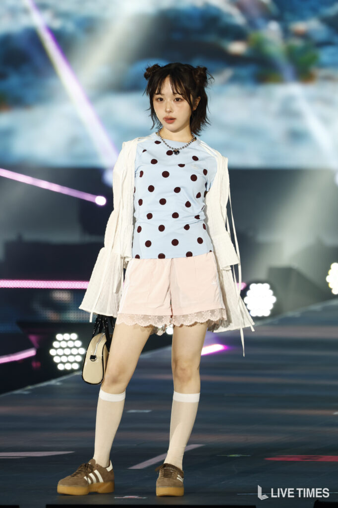 Young woman on stage wearing a light blue polka-dot shirt, pink shorts, white cardigan, knee-high socks, and brown sneakers, carrying a beige bag with lights/stage backdrop behind.