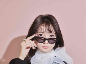 Portrait of a woman with dark hair wearing black sunglasses and a sheer white scarf against a pale pink background.