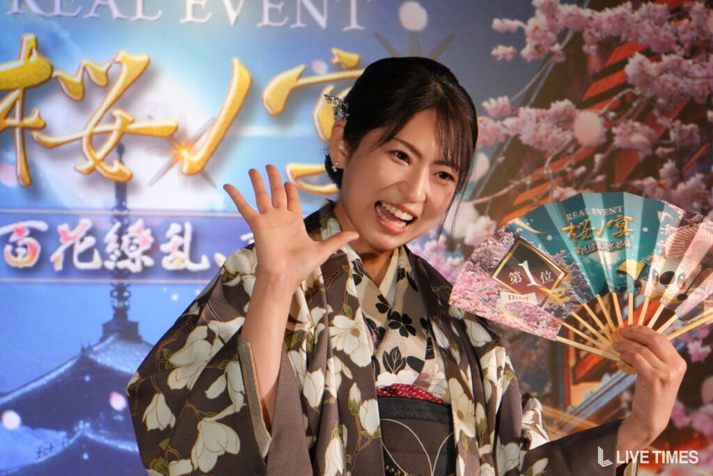 Smiling woman in a floral kimono waves to the camera while holding a decorative fan against a festival backdrop of pink blossoms and banners.