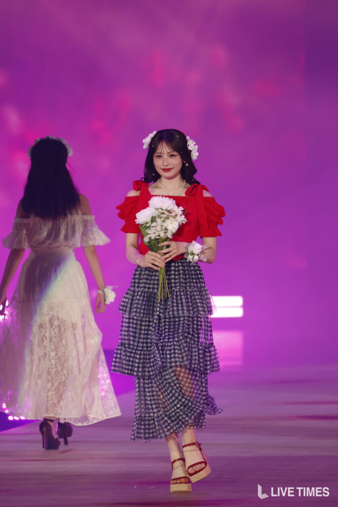 Young woman in a red off-shoulder top and tiered plaid skirt holding a bouquet on a pink-lit stage, with flowers in her hair.