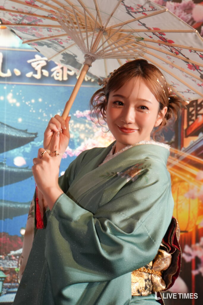 Smiling woman in a green kimono holds a floral parasol with cherry blossom design.