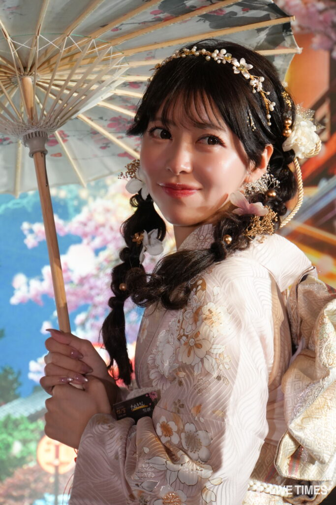 Young woman in a pale floral kimono holding a decorative parasol, smiling gently under a floral backdrop.