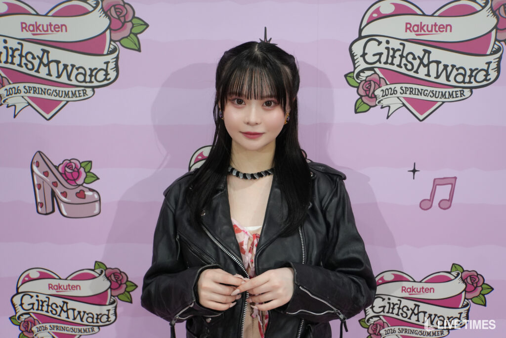 Young woman posing in front of a pink backdrop with GirlsAward logos and Rakuten branding, wearing a black leather jacket and necklace.