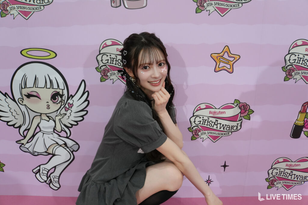 Smiling young woman posing in a gray dress against a pastel pink 'GirlsAward' backdrop with cartoon angels and logos.