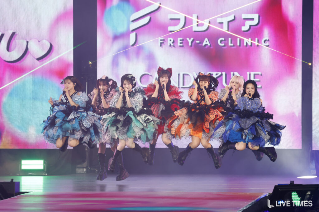 Nine female performers in colorful, layered dresses jump mid-performance on a bright stage backdrop.