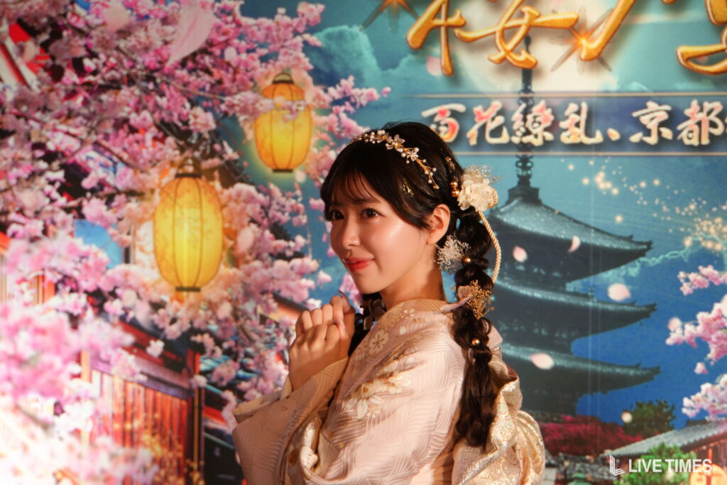 Portrait of a woman in a pale floral kimono with ornate hair accessories, posing in front of cherry blossoms and lanterns.