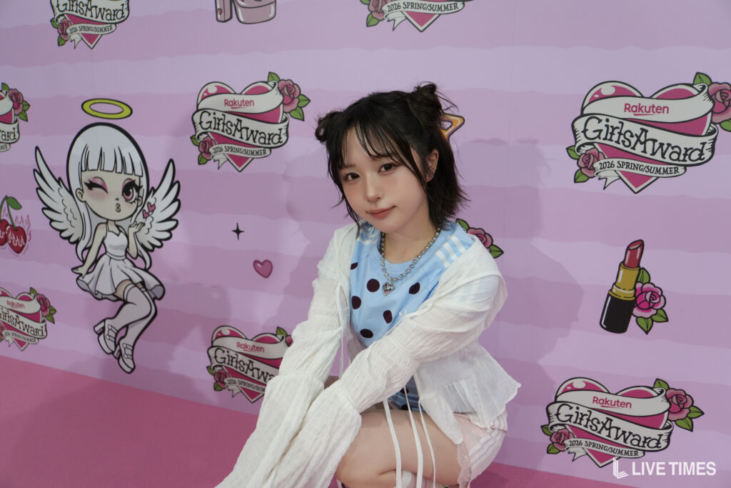 Young woman posing and smiling slightly in front of a pink GirlsAward backdrop, wearing a blue polka-dot top and white cardigan.