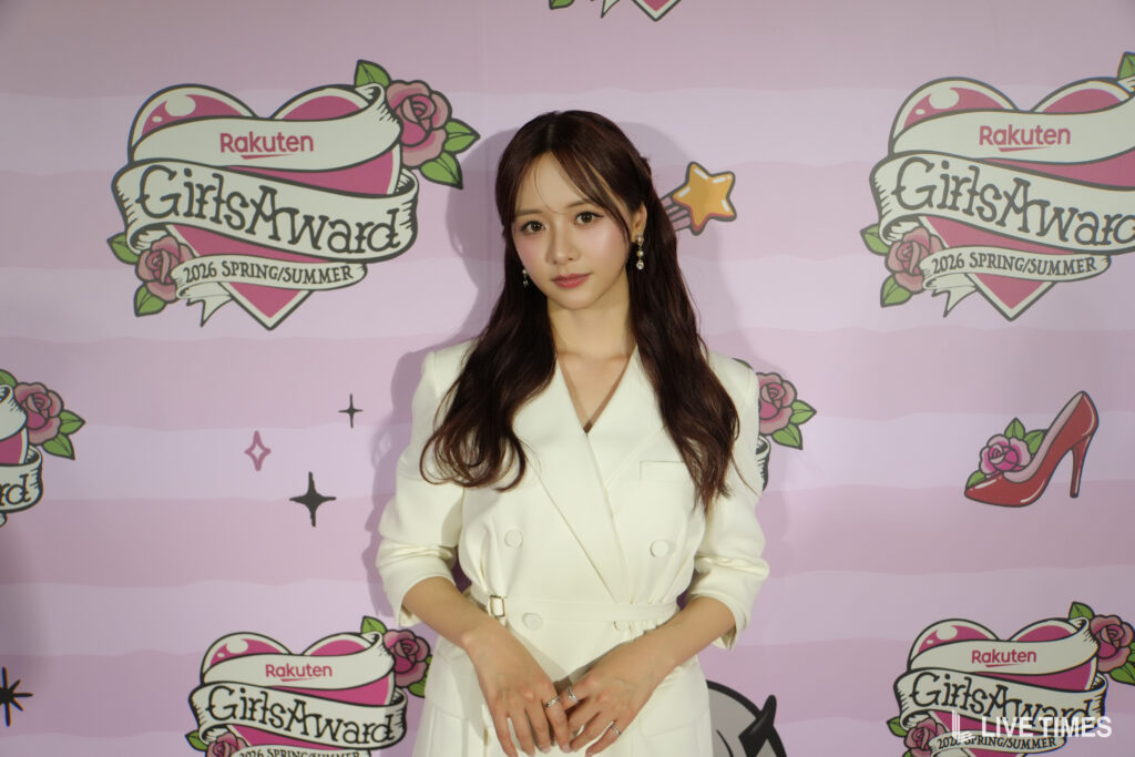 Woman in a white blazer posing in front of a pink backdrop with 'GirlsAward' and Rakuten logos at a fashion event.