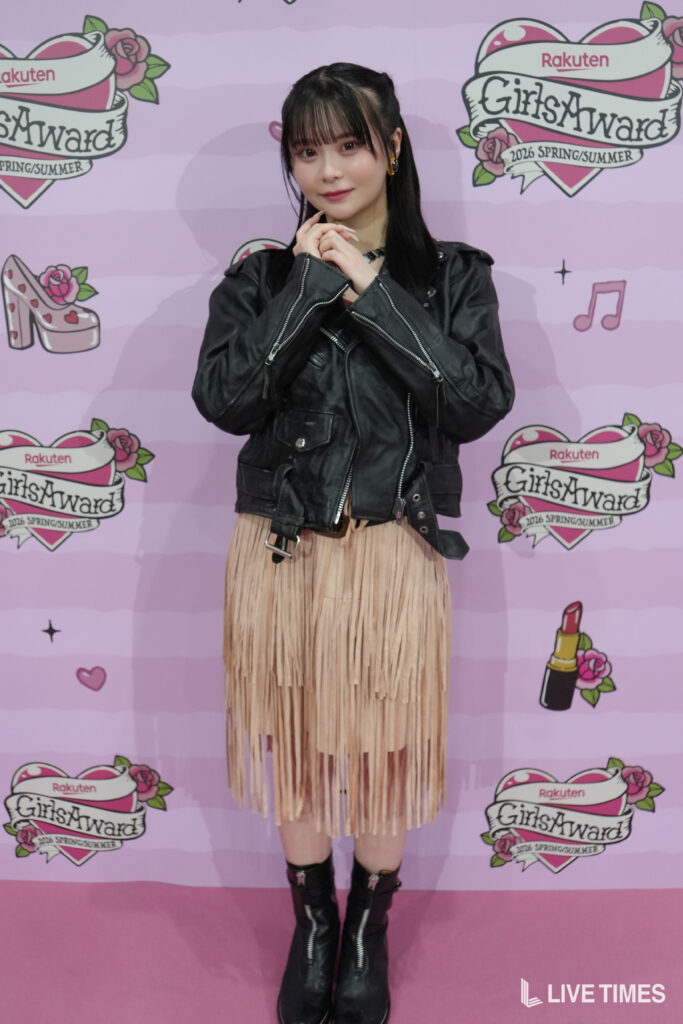 Young woman posing for a photo in a pink backdrop with GirlsAward logos, wearing a black leather jacket, beige fringe skirt, and black boots.