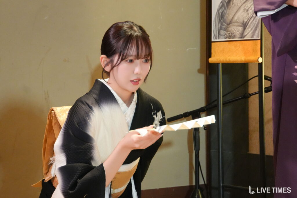 Young woman in a black-and-gold kimono reading from a decorative paper on a stage.