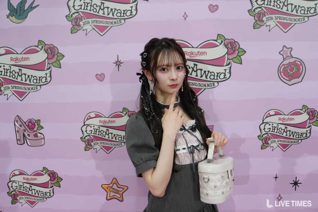 Young woman posing in front of a pink GirlsAward backdrop, wearing a gray dress and holding a white handbag.
