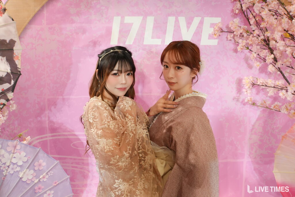 Two women in pink lace dresses posing together against a pink cherry-blossom backdrop, with a bold 'LIVE' sign in the background.