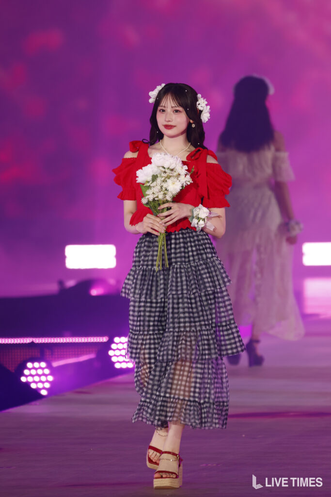 Young woman walking a runway in a red off-shoulder top and layered gingham skirt, holding a bouquet of white flowers.