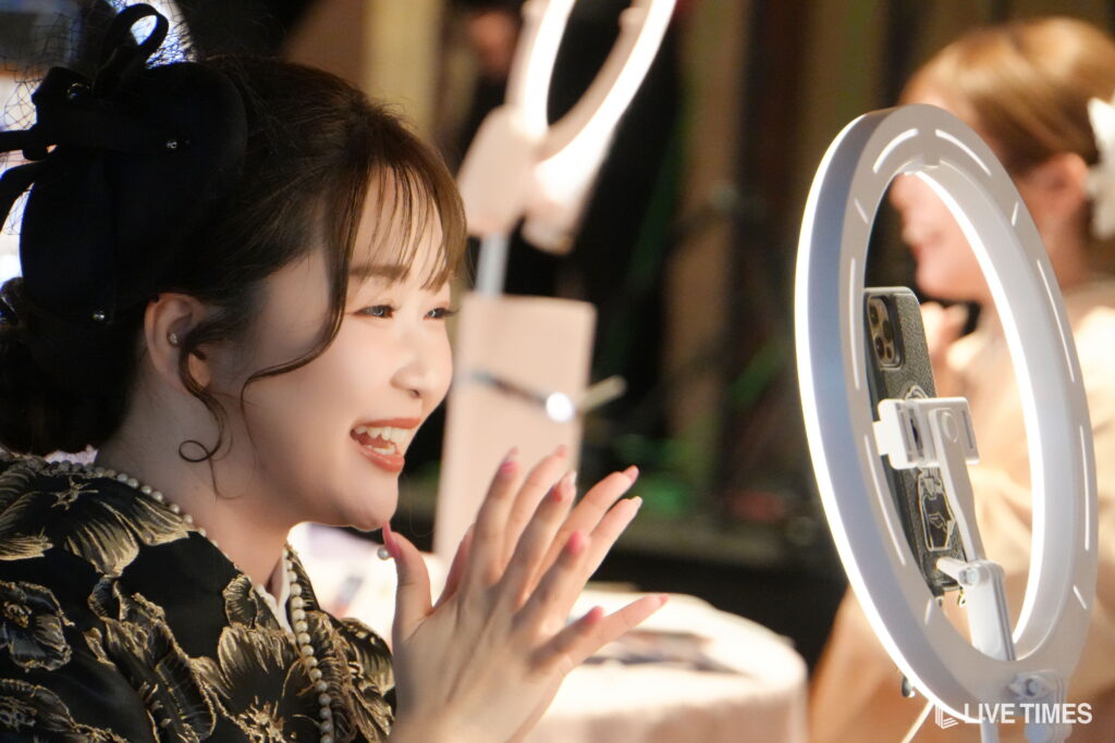 Smiling woman in a black and gold dress claps beside a circular ring light at a live event.