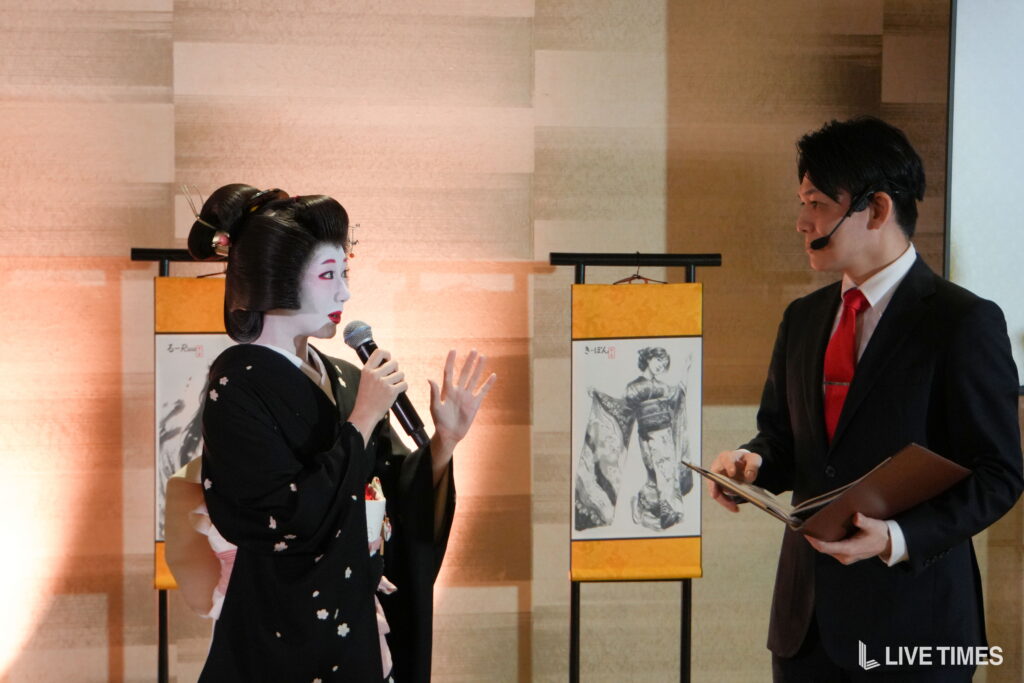 Geisha in a kimono speaking into a microphone while a suited man with a headset listens on stage.
