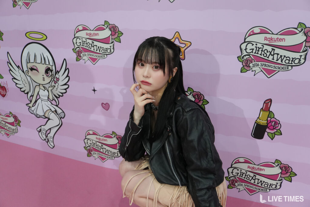 Young woman with long dark hair in a black leather jacket poses in front of a pink GirlsAward backdrop with logos and decorations.
