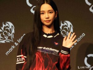 Young woman with long black hair waving at the camera on a stage backdrop with decorative graphics.