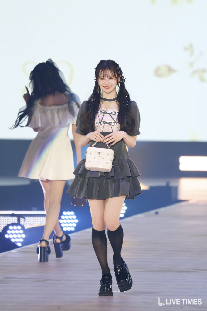 Model on a runway wearing a dark layered dress, black socks, shiny shoes, and a small white handbag, smiling at the camera in a stage setting.