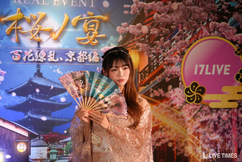 Young woman in a lace dress holding colorful decorative fans on a stage with cherry blossoms and a 17LIVE backdrop/logo visible in the corner.