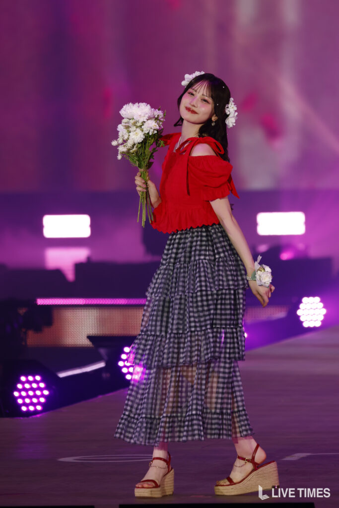 Performer on stage in a red off-shoulder top and a layered checkered skirt, holding a bouquet of white flowers, wearing a flower crown under purple stage lights.