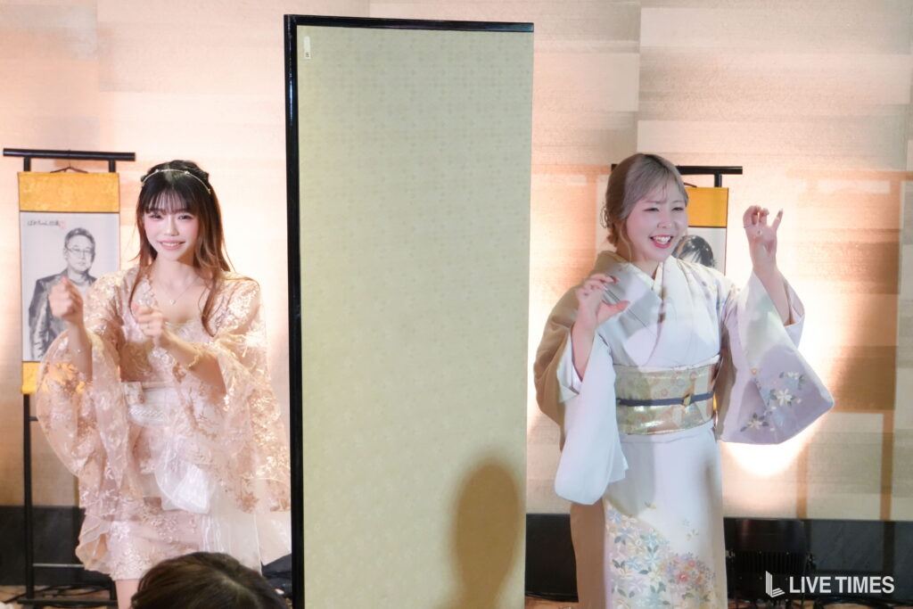 Two women in kimono performing on a stage with beige folding screens, smiling and gesturing gracefully.