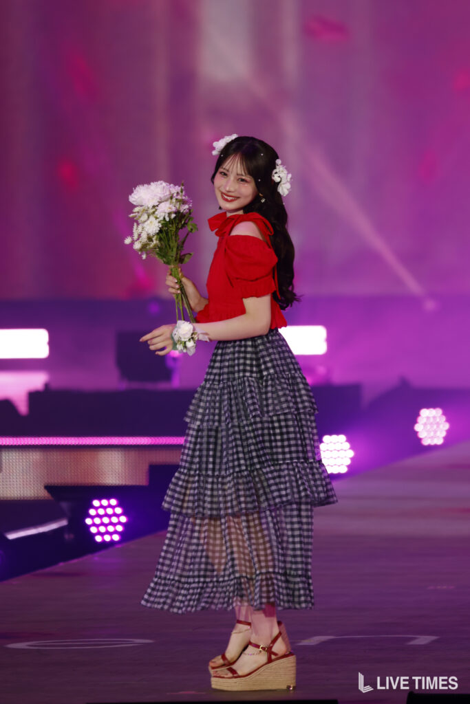Young woman on stage in a red off-shoulder top and tiered checkered skirt, holding a bouquet and smiling amid pink-purple stage lights.