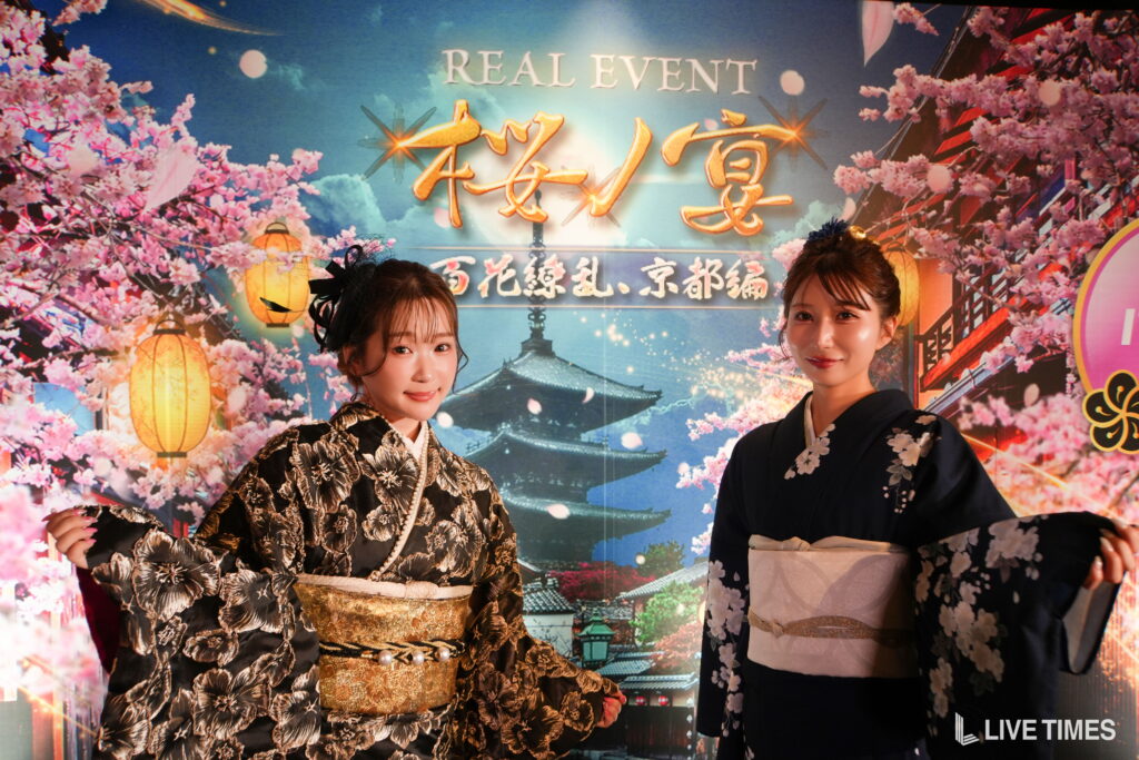 Two women in ornate kimonos posing in front of a cherry-blossom event backdrop with lanterns and 'REAL EVENT' signage.