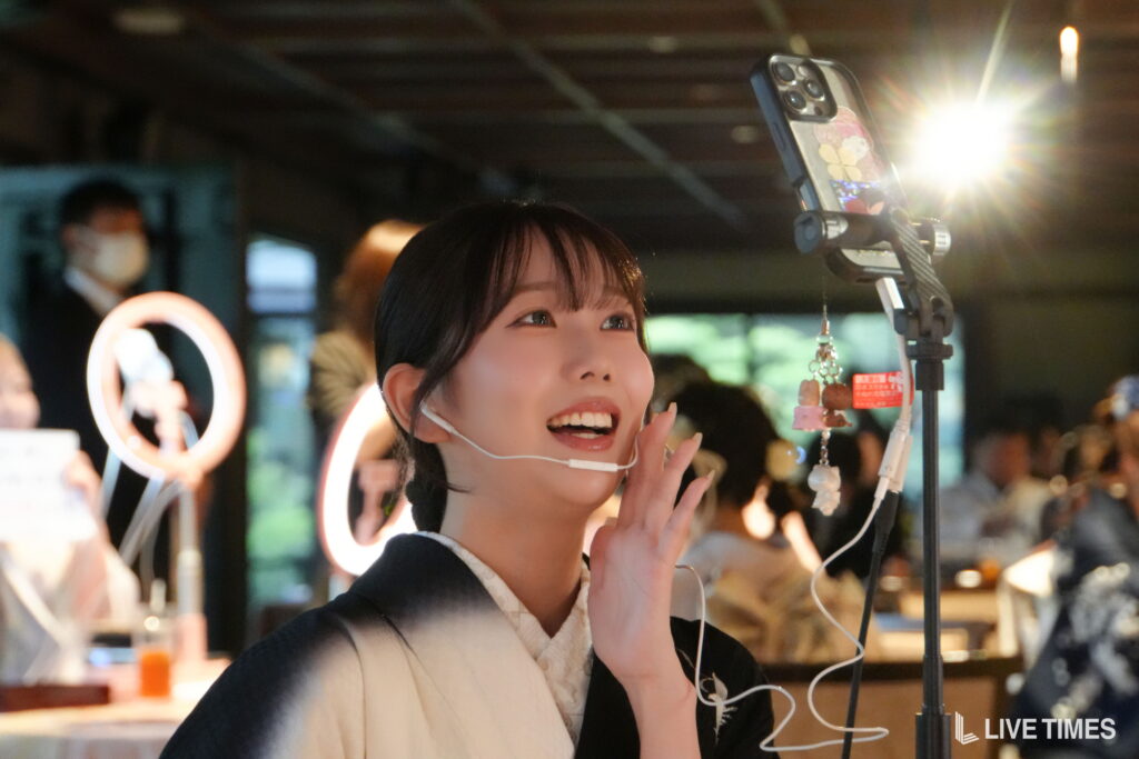 Young woman wearing a headset microphone, smiling, singing into a microphone with ring light in the background in a busy indoor setting.