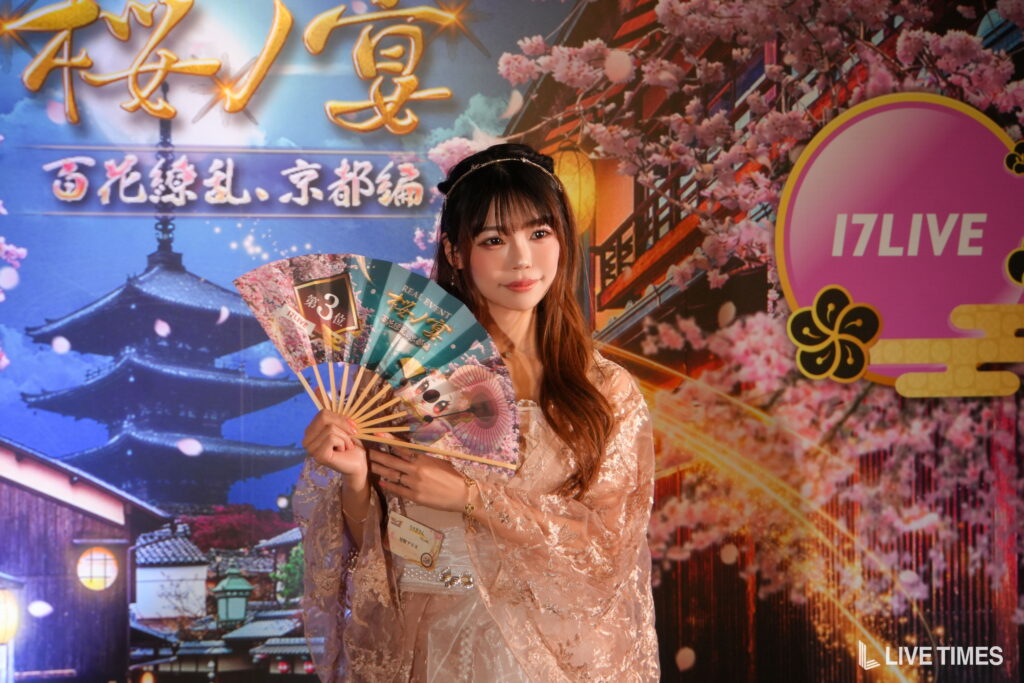 Young woman in a lace dress with a headband, holding colorful folding fans, posing in front of a cherry-blossom stage backdrop with a 17LIVE logo.