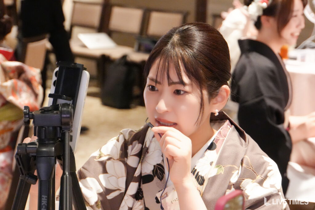 Woman in a floral kimono adjusting a camera on a tripod at an indoor event.