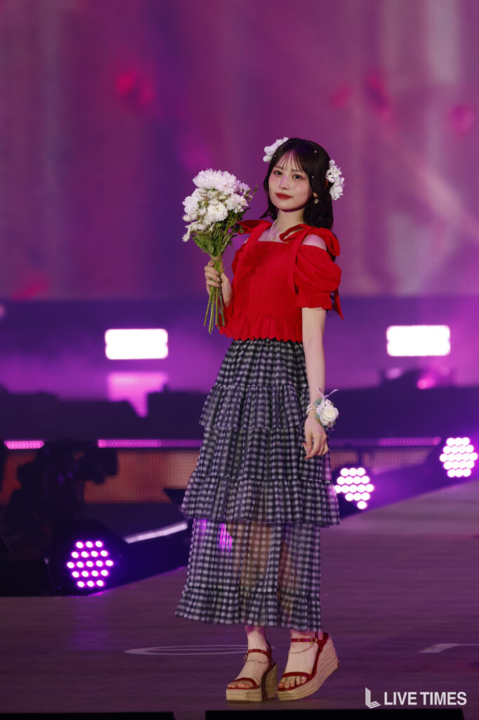 Performer on stage in a red off-shoulder top and tiered gingham skirt, holding a bouquet of white flowers with floral accessories in her hair.