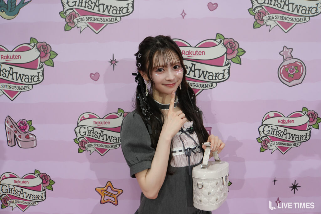 Young woman posing with her finger to her lips in front of a pink GirlsAward backdrop with Rakuten branding and heart decorations.